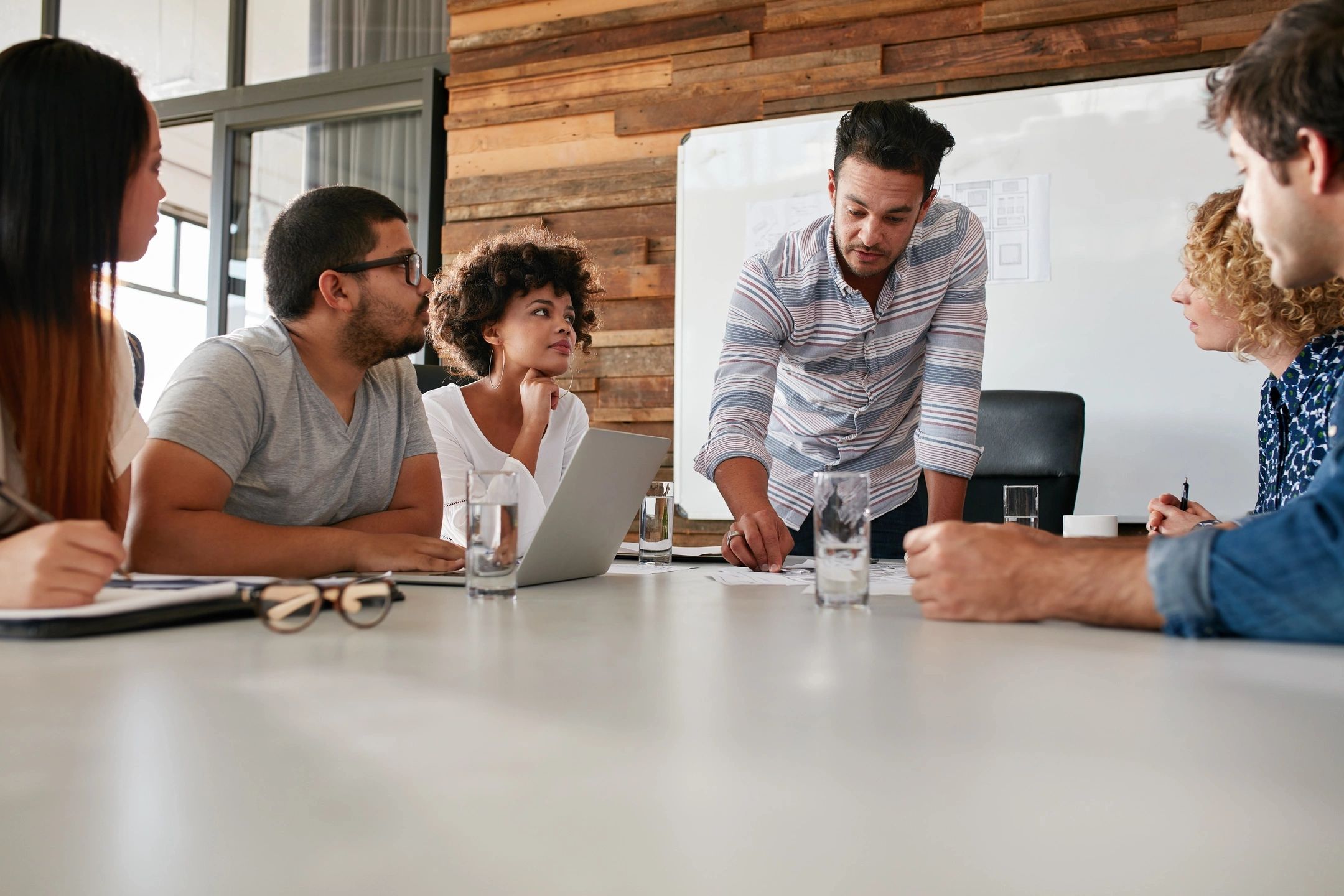 A group of people sitting in a meeting. One guy is standing and taking notes at the end of the table.